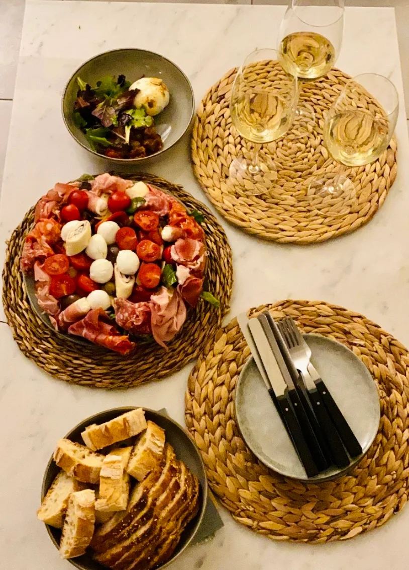 Table dressée : plateau de charcuteries italiennes et mozzarella, corbeille de pain, trois verres de vin blanc sur le marbre.