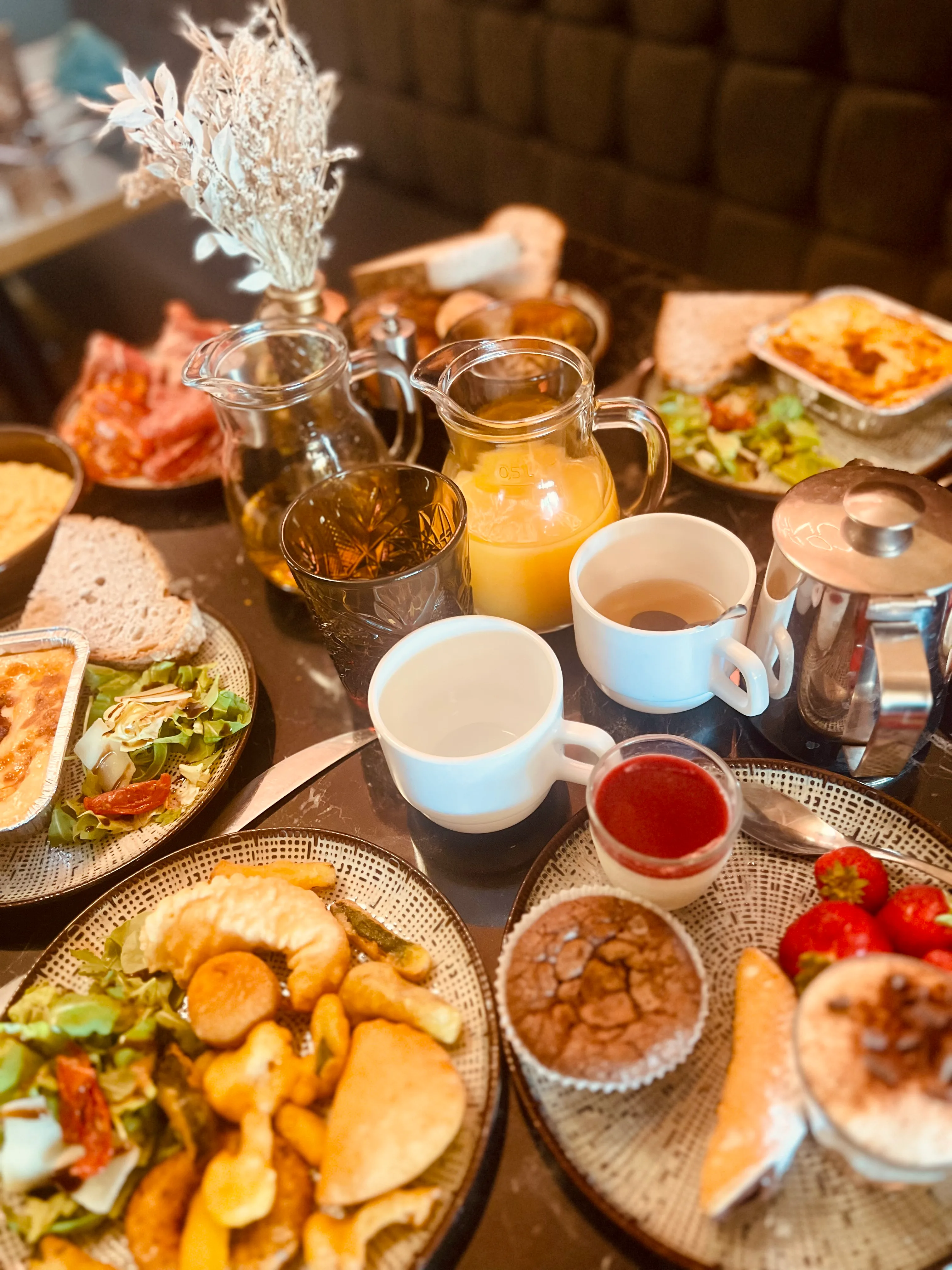 La table du brunch du dimanche : charcuteries, lasagnes, fritures, pain, jus d'orange, théière et pâtisseries maison.