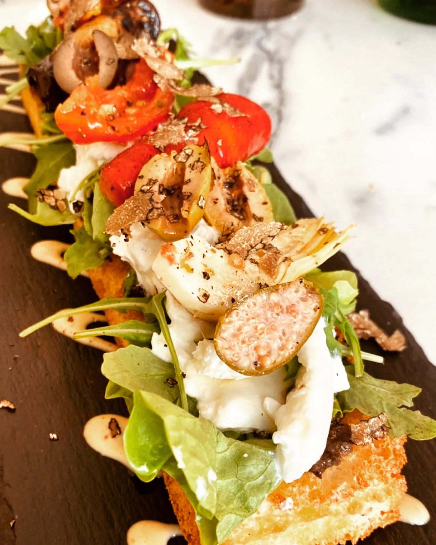 Focaccia grillée en bouchées sur ardoise : burrata, roquette, tomates confites, artichauts, figue et copeaux de truffe noire.